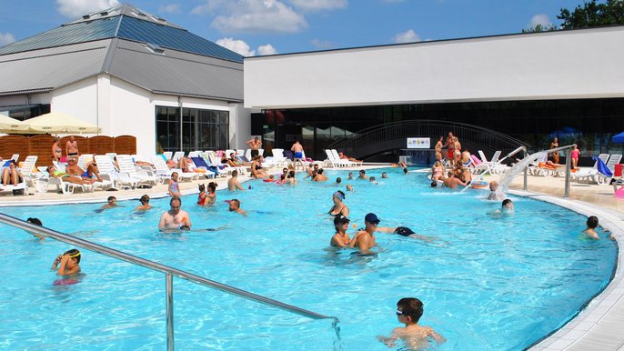 Zewnętrzny basen wypływowy w słoneczny dzień wypełniony Falowiczami, którzy relaksują się w błękitnej wodzie lub na leżakach wokół basenu. W tle hala basenowa z basenem sportowym. 