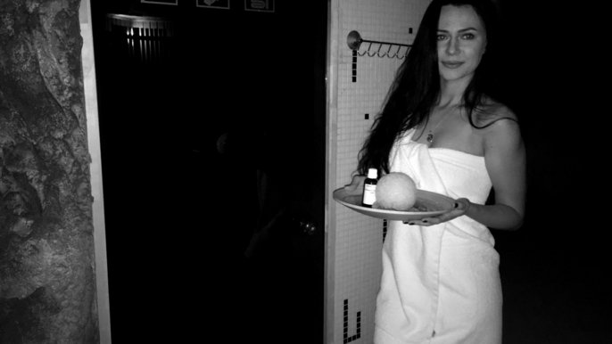 Zdjęcie czarno-białe. Sauanmistrzyni Sylwia Skarbek. Młoda kobieta z ciemnymi włosami owinięta białym ręcznikiem trzyma w dłoniach tacę z lodową kulą i olejkiem eterycznym i lekko się uśmiecha. 