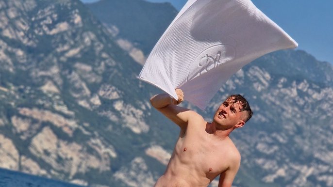 Mężczyzna bez koszulki machający białym ręcznikiem na tle gór. Ujęcie obejmuje mężczyznę od pasa w górę. 