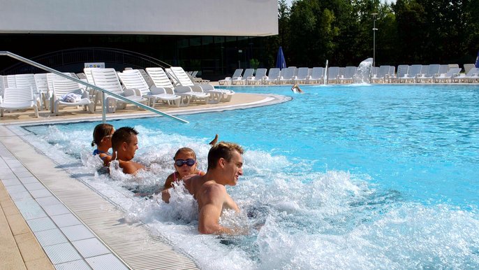 Czteroosobowa rodzina relaksuje się w basenie wypływowym w słoneczny dzień. Rodzice wraz z kilkuletnią córką i nastoletnim synem korzystają z powietrznych leżaków przy brzegu basenu. W tle hala z basenem sportowym. 