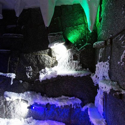 Wnętrze groty śnieżnej - ciemne kamienie na ścianach, warstwa śniegu na podłodze, wiszące stalaktyty z sufitu pokryte są śniegiem. Resztki śniegu znajdują się również na ścianach. Oświetlenie w kolorze zielonym i niebieskim. 