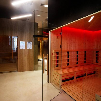 Zbliżenie na saunę infrared obudowaną z zewnątrz ciemnymi płytkami, wewnątrz zapalone jest czerwone światło i ściany oraz podłoga zabudowane są drewnem. W tle widać saunę z muzykoterapią i tężnię solną. 