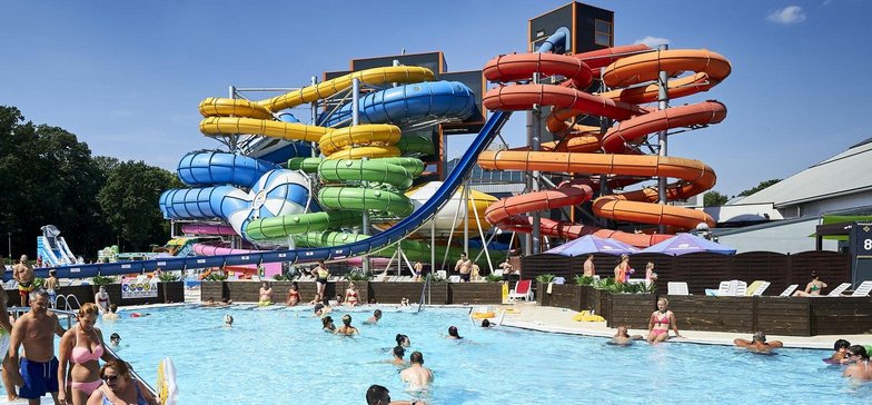 Zewnętrzny basen wypływowy w słoneczny dzień wypełniony ludźmi. W tle wieże z kolorowymi MegaZjeżdżalniami.