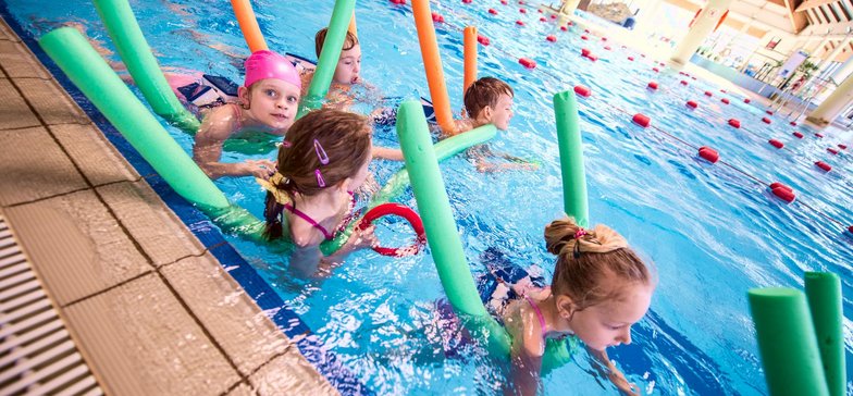 Grupa dzieci przy brzegu wewnętrznego basenu sportowego. Każde z nich korzysta z kolorowych makaronów pływackich.