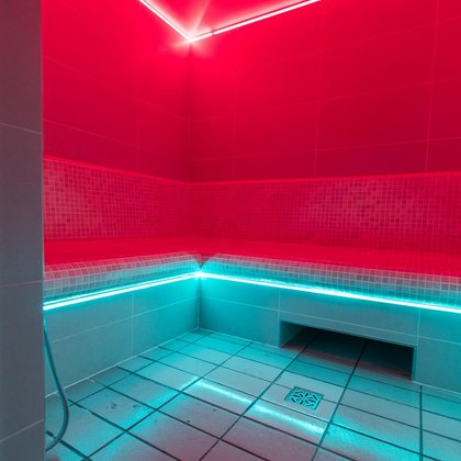 Wnętrze sauny parowej oświetlone na czerwono. Wzdłuż ścian dostępne są ceramiczne siedziska podświetlone kolorem turkusowym. 