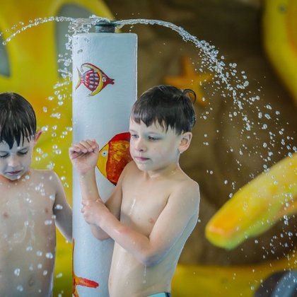 Dwóch kilkuletnich chłopców bawi się pod niewielką fontanną ozdobioną malunkami kolorowych rybek. W tle widać żółtą Ośmiornicę Matyldę. 