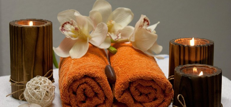 Pomarańczowe ręczniki zwinięte w rulon otoczone ciemnymi świecami i jasnymi kwiatami.