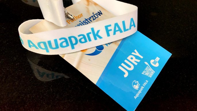 Beżowo-niebieska kartonowa wejściówka z logiem turnieju PTS Freestyle i napisem "Jury" zawieszona na białej smyczy reklamowej z logotypem Aquaparku FALA. 