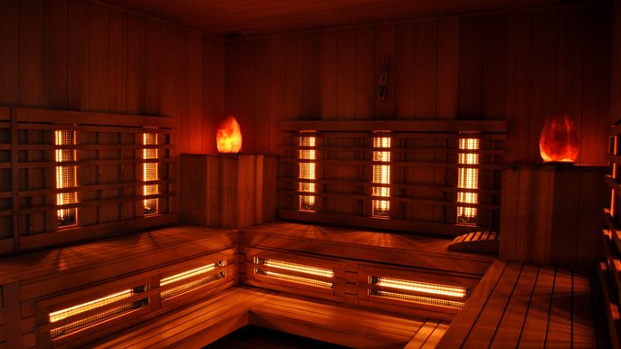 Wnętrze sauny na podczerwień. Widać trzy ściany wzdłuż, których rozmieszczone są siedziska i promienniki podczerwone oświetlające saunę pomarańczowym światłem. W dwóch rogach sauny dodatkowo funkcjonują lampy w kształcie kryształów. 