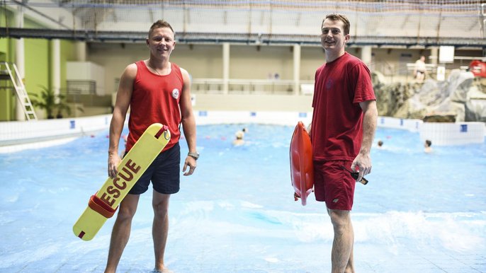 Dwóch ratowników w czerwonych strojach WOPR stoi przy basenie z falą. Ratownik po lewo trzyma żółtą rzutkę ratowniczą, mężczyzna po prawo pomarańczową bojkę. 