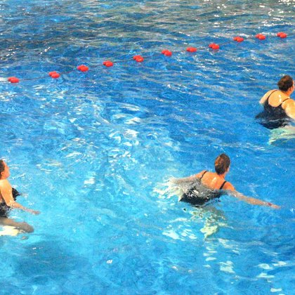 Zbliżenie na panie ćwiczące w basenie z efektem fali - lekko pochylone, wyciągają w bok jedną z rąk. 