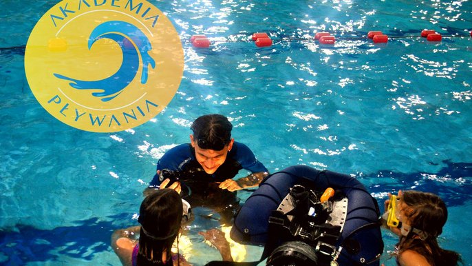 Jeden z torów w basenie sportowym. Instruktor w niebieskim uniformie przekazuje dwójce dzieci instrukcje jak prawidłowo korzystać z butli z aparatem do snurkowania. W lewym górnym rogu żółto-niebieskie logo Akademii Pływania. 