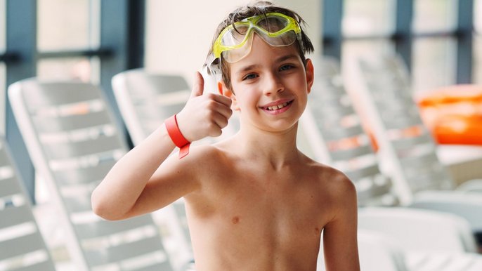 Uśmiechnięty kilkuletni chłopiec unosi w górę kciuk prawej ręki. Na czoło zsunięte ma żółte okularki pływackie. 