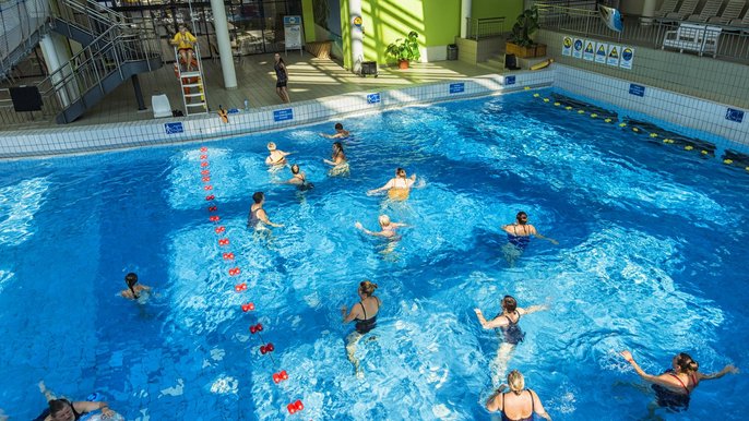 Basen z efektem fali - efekt jest wyłączony, w głębszej części basenu odbywają się zajęcia zorganizowane. Kilkunastoosobowa grupa w wodzie powtarza ćwiczenia instruktorki stojącej na brzegu. 