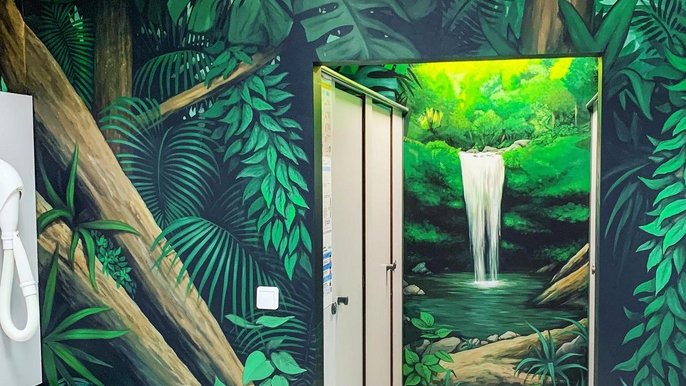 Szatnia Strefy Saun: widać boksy przebieralni, a na ścianach wokół mural imitujący rajską dżunglę z wodospadem i drzewami z zielonymi liśćmi w różnych kształtach i odcieniach. 