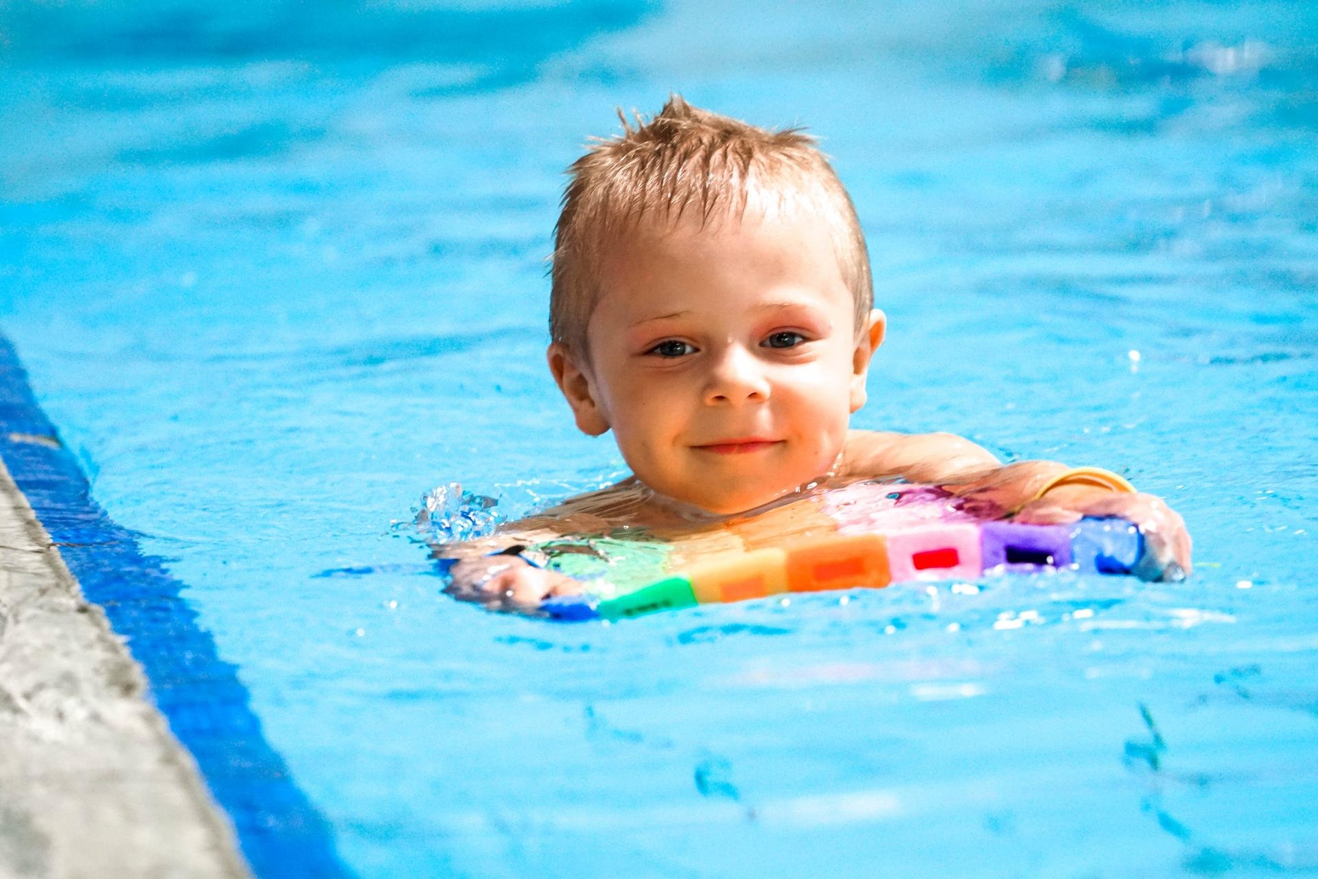 Uśmiechnięty kilkuletni chłopiec na jednym z torów basenu sportowego. Płynie na brzuchu przed sobą trzymając kolorową deskę do pływania. 