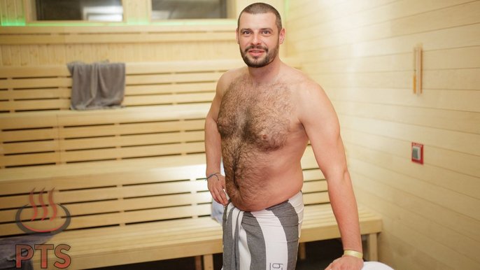 Saunamistrz Łukasz Gapiński: mężczyzna w ciemnych włosach, ubrany w ręcznik. Pozuje w saunie, na tle jasnych drewnianych ławek. 