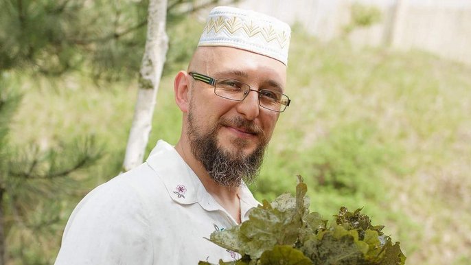 Saunamistrz Marek Olszewski - mężczyzna w średnim wieku w okularach ma na sobie biały strój i trzyma w dłoniach pęk liści dębu. 