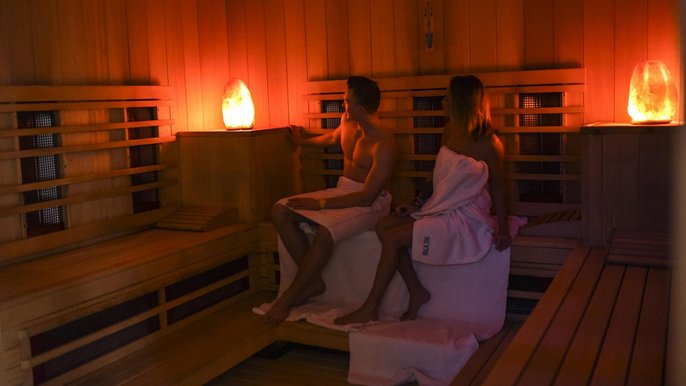 Dwoje młodych ludzi w saunie infrared. Oboje owinięci są białymi ręcznikami, ręczniki rozłożone są też na drewnianych ławach sauny, spoglądają na klimatyczną lampę rzucającą pomarańczowe światło w saunie. 