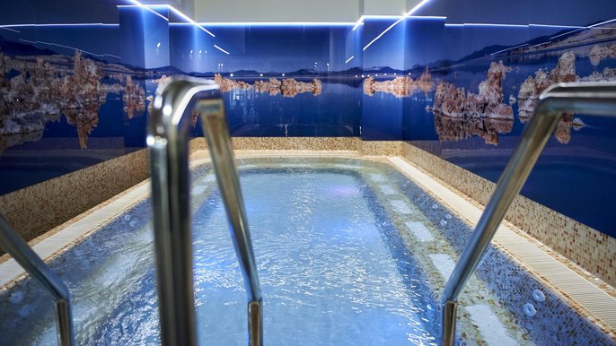 Basen solankowy z solą z Morza Martwego. Ujęcie z perspektywy schodów prowadzących do basenu. Błękitna woda, dookoła krawędź obudowana beżową mozaiką, a na ścianach wokół fototapeta za szkłem ukazująca panoramę Morza Martwego. 