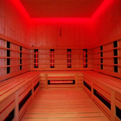 Wnętrze sauny infrared - podświetlone w kolorze czerwonym. Ściany i podłoga obudowane drewnem. Na ścianach i pod ławą ciągnącą się wzdłuż trzech ścian widać promienniki podczerwone. 