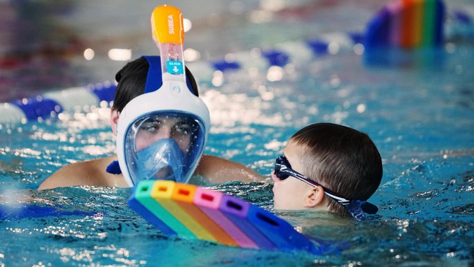 Nastolatek w masce do snorkelingu asekuruje kilkuletniego chłopca w basenie sportowym. Młodszy chłopiec trzyma się kolorowej deski pływackiej i ma na sobie ciemne okulary pływackie. 
