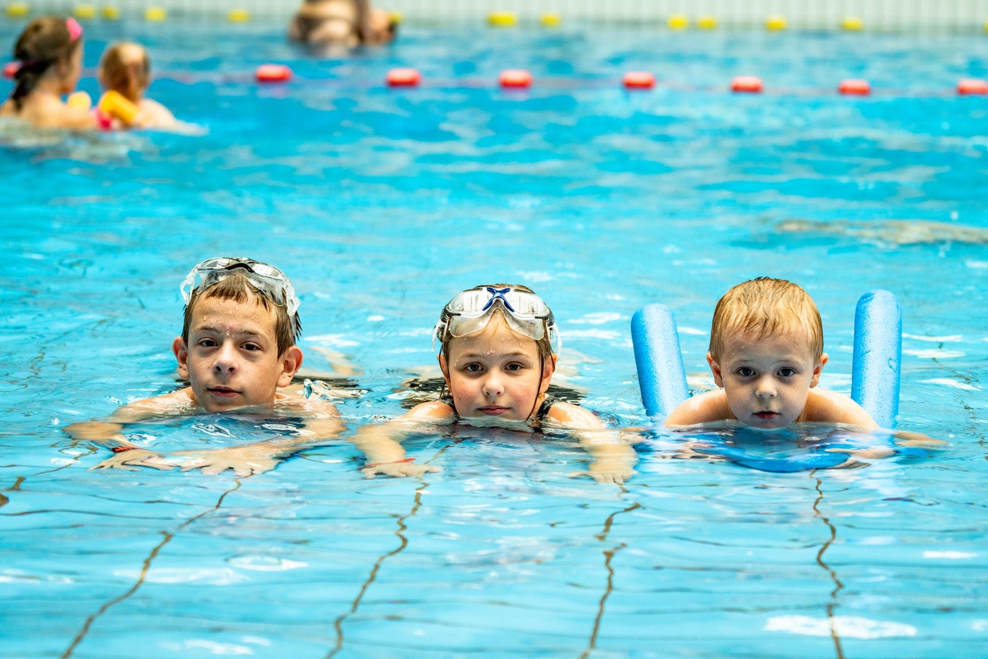 Troje dzieci unosi się na wodzie w płytszej części basenu z efektem fali. Od lewej nastoletni chłopiec z okularkami pływackimi na głowie, w środku dziewczynka, po prawo kilkuletni chłopiec z niebieskim makaronem pływackim. 
