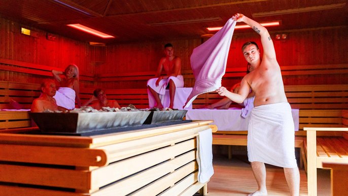 Zdjęcie wewnątrz sauny fińskiej podczas ceremonii saunowej, nad piecem saunamistrz macha ręcznikiem, w tle klienci rozkoszujący się seansem siedzą na drewnianych ławach sauny. 