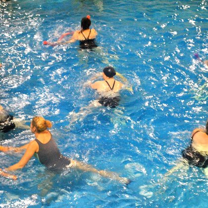Zbliżenie na grupę ćwiczącą w basenie. Panie mają w dłoniach wygięte, kolorowe wodne makarony i pochylają się z nimi w jedną ze stron. 