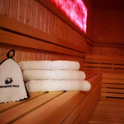 Wnętrze sauny solnej - zbliżenie na czapkę saunową z logotypem Aquaparku FALA i trzy białe ręczniki złożone na drewnianej ławie. W tle widać panel solny podświetlony w kolorze czerwonym i dwa rzędy drewnianych siedzisk. 