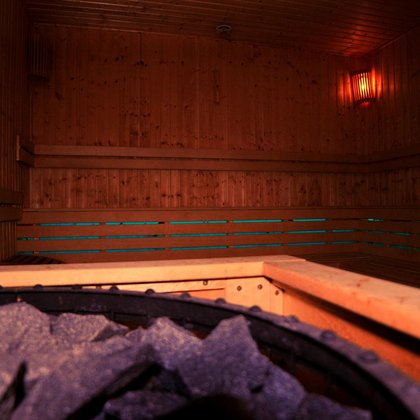 Wnętrze sauny z muzykoterapią z perspektywy pieca saunowego. Na pierwszym planie ciemne kamienie, a w tle drewniane ławy wzdłuż dwóch ścian podświetlone w kolorze błękitnym. 