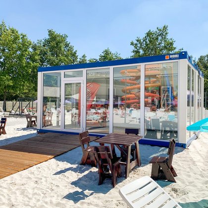 Przeszklony bar znajdujący się na piaszczystej plaży. W szybach punktu gastronomicznego odbijają się MegaZjeżdżalnie, wokół rozstawione są drewniane stoliki z krzesełkami i leżaki. 