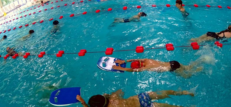 Grupa dzieci w wewnętrznym basenie sportowym. Płynące stylem klasycznym trzymają przed sobą deski do nauki pływania.