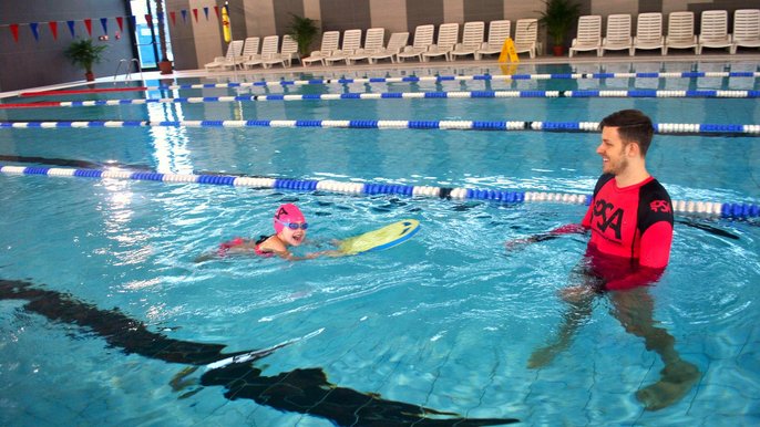 Basen sportowy. Na jednym z torów instruktor w różowej koszulce PSA nadzoruje ćwiczenia wykonywane przez kilkuletnią dziewczynkę w różowym czepku pływackim i z żółtą deską pływacką przed sobą. 