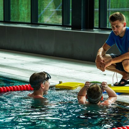 Instruktor pływania kuca przy brzegu basenu sportowego i rozmawia z dwiema dziewczynkami w basenie, przekazuje instrukcje do dalszych ćwiczeń. 
