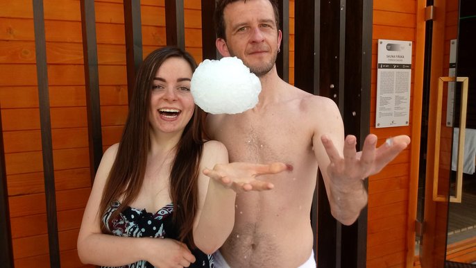 Saunamistrzowie Sylwia Drobiszewska i Emil Tyrolczyk pozują uśmiechnięci podrzucając lodowe kule. W tle charakterystyczna drewniana obudowa dużej sauny zewnętrznej. 