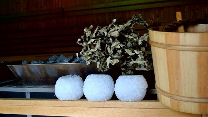 Zbliżenie na trzy lodowe kule, drewniane wiadro na wodę niezbędną podczas seansu saunowego oraz brzozowe witki. Wszystko to zostało ułożone na drewnianej obudowie pieca saunowego. 
