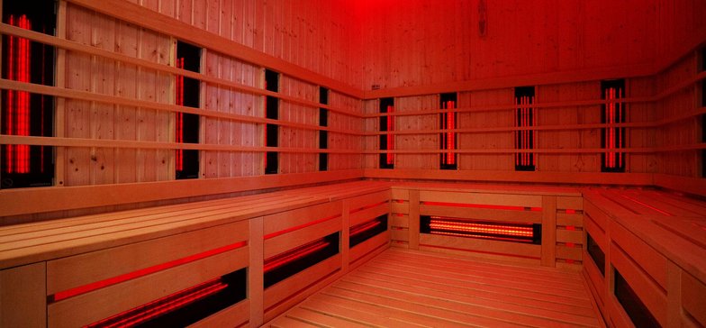 Wnętrze sauny infrared - podświetlone w kolorze czerwonym. Ściany i podłoga obudowane drewnem. Na ścianach i pod ławą ciągnącą się wzdłuż trzech ścian widać promienniki podczerwone.