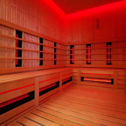 Wnętrze sauny infrared - podświetlone w kolorze czerwonym. Ściany i podłoga obudowane drewnem. Na ścianach i pod ławą ciągnącą się wzdłuż trzech ścian widać promienniki podczerwone. 