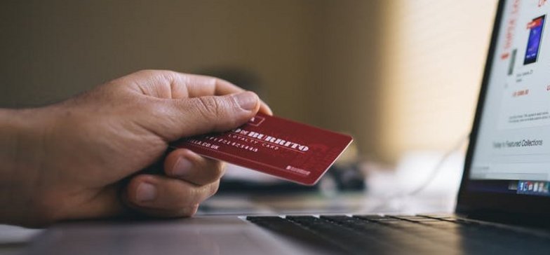 Zbliżenie na laptopa - widać fragment klawiatury i monitora. Przy komputerze dłoń trzymająca czerwoną kartę kredytową.