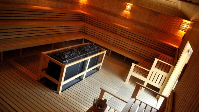 Zdjęcie wnętrza dużej sauny zewnętrznej w Strefie Saun. W centralnym punkcie piec z ciemnymi kamieniami, podłoga i ściany wyłożone drewnem, wokół ścian trzy rzędy drewnianych ław. 