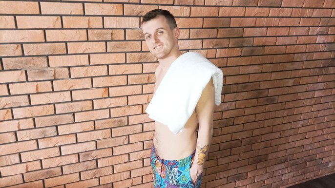 Marcin Sikorski, mężczyzna bez koszulki, z białym ręcznikiem na ramieniu. Mężczyzna ubrany jest w kolorowe spodenki. Pozuje na tle ściany wykonanej z czerwonych cegieł. Na lewym nadgarstku ma kolorowy tatuaż – miś z czerwoną koka 