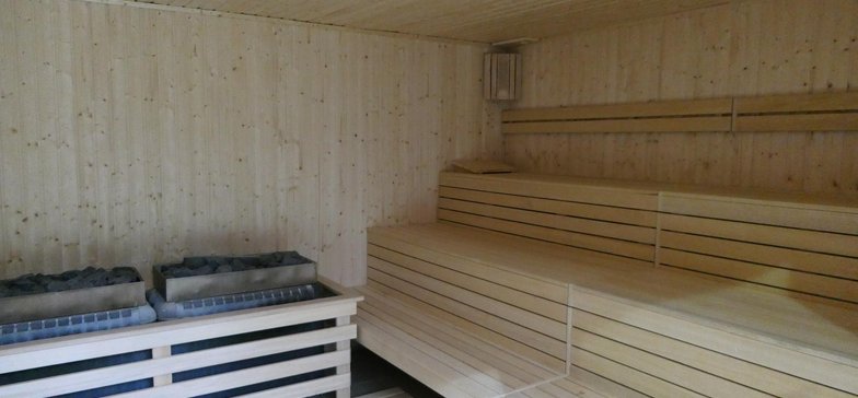 Wnętrze małej sauny zewnętrznej. Cała sauna obudowana drewnem. Po prawej trzy rzędy drewnianych siedzisk, przy ścianie naprzeciw podwójny, prostokątny piec pokryty kamieniami.