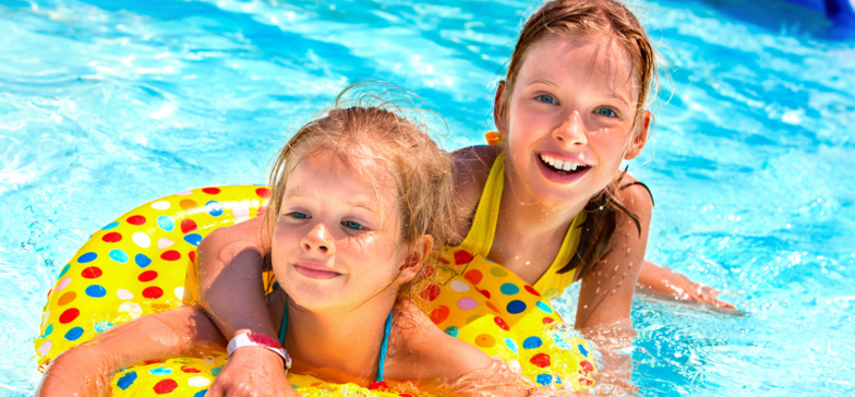 Dwie uśmiechnięte rudowłose dziewczynki pozują w basenie. Dziecko na pierwszym planie jest w dmuchanym kole do pływania. 