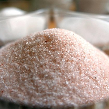 Sól peelingowa o lekko różowym zabarwieniu wsypana jest do szklanej miski, w tle widać zarys innych naczyń z białymi granulkami. 