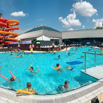 Całoroczny basen wypływowy wypełniony kąpiącymi się ludźmi. Po prawo w tle widać halę basenową z logotypem Aquaparku, a po lewo kolorowe MegaZjeżdżalnie i Zjeżdżalnię Kamikaze. 