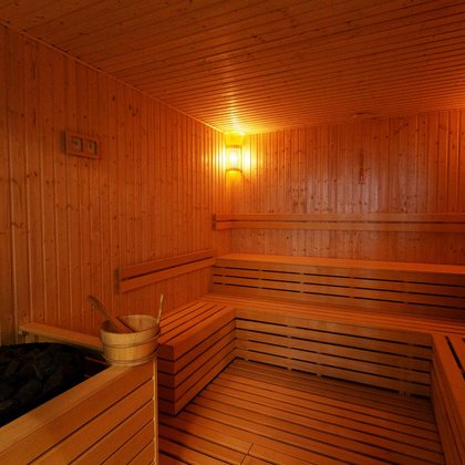 Wnętrze sauny z muzykoterapią. Podłoga i ściany zabudowane drewnem, wzdłuż dwóch ścian drewniane ławy z trzema rzędami siedzisk, w prawym dolnym rogu okrągły piec z ciemnymi kamieniami. 