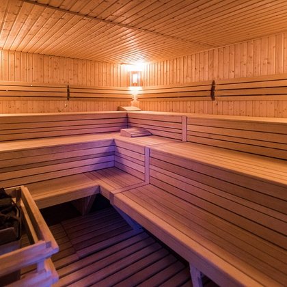 Wnętrze sauny fińskiej wewnętrznej. W lewym dolnym rogu widać piec z rozłożonymi kamieniami, a wzdłuż dwóch ścian sauny drewniane ławy z trzema rzędami siedzisk. 