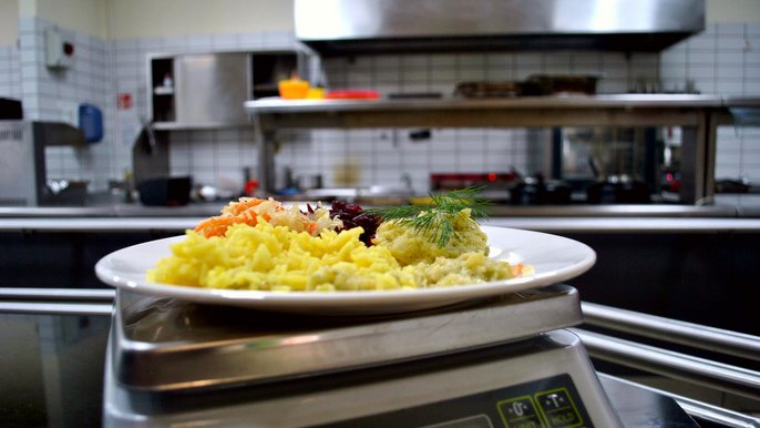 Zbliżenie na talerz wypełniony ryżem z curry, kolorowymi surówkami i mięsem w jasnym sosie. Talerz umieszczony jest na wadze, a w tle widać otwartą część kuchni restauracji. 