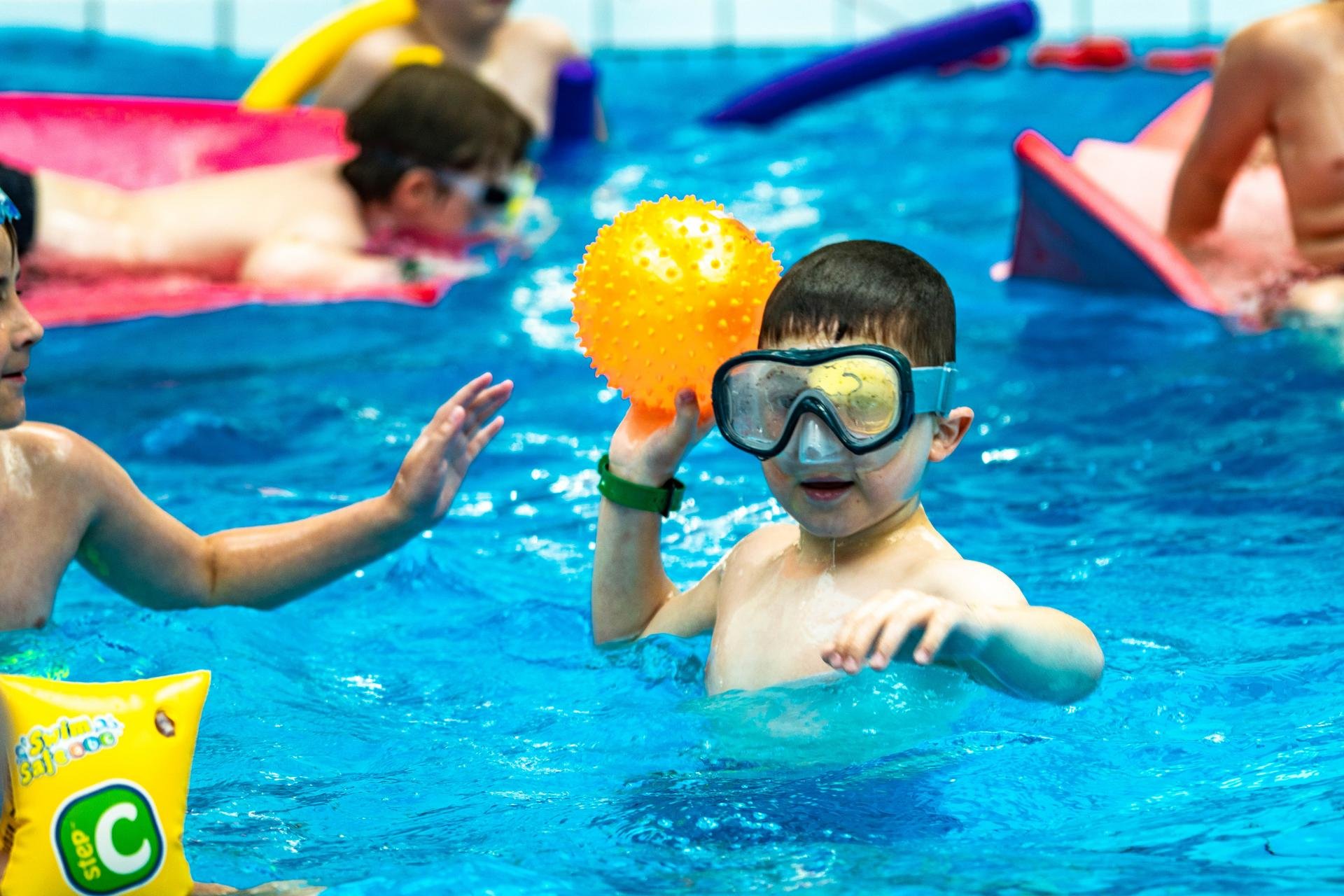 Dzieci w basenie bawią się z wykorzystaniem gumowych piłek, piankowych materacy i makaronów do pływania. 